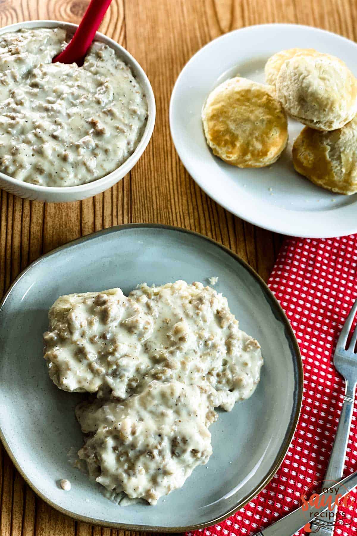 Sausage gravy over three biscuits with a plate of biscuitsand bowl of gravy  to the side