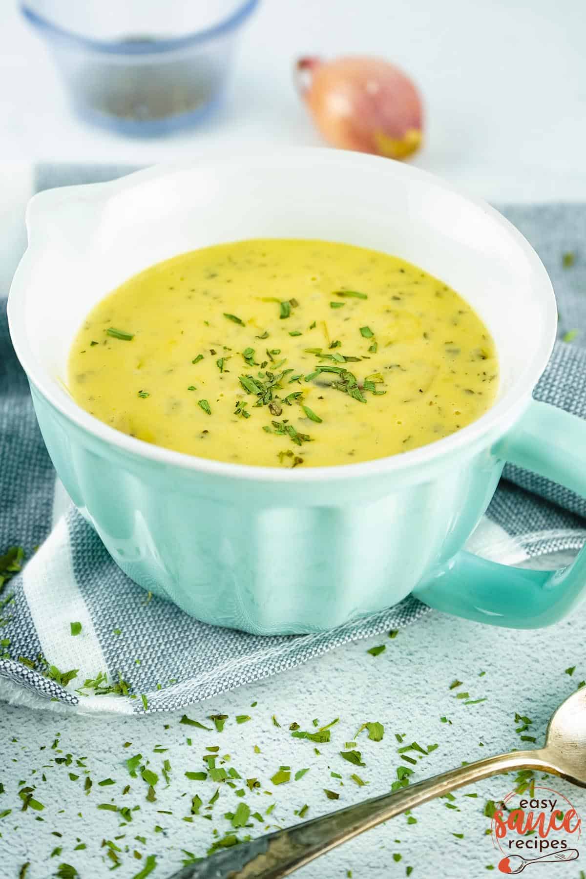 a turquoise gravy boat filled with bearnaise sauce resting on a grey towel, with a shallot, spoon and herbs in the background