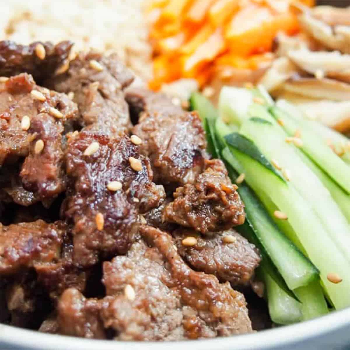 close up of bulgogi beef in a bowl next to sliced cucumbers with sesame seeds