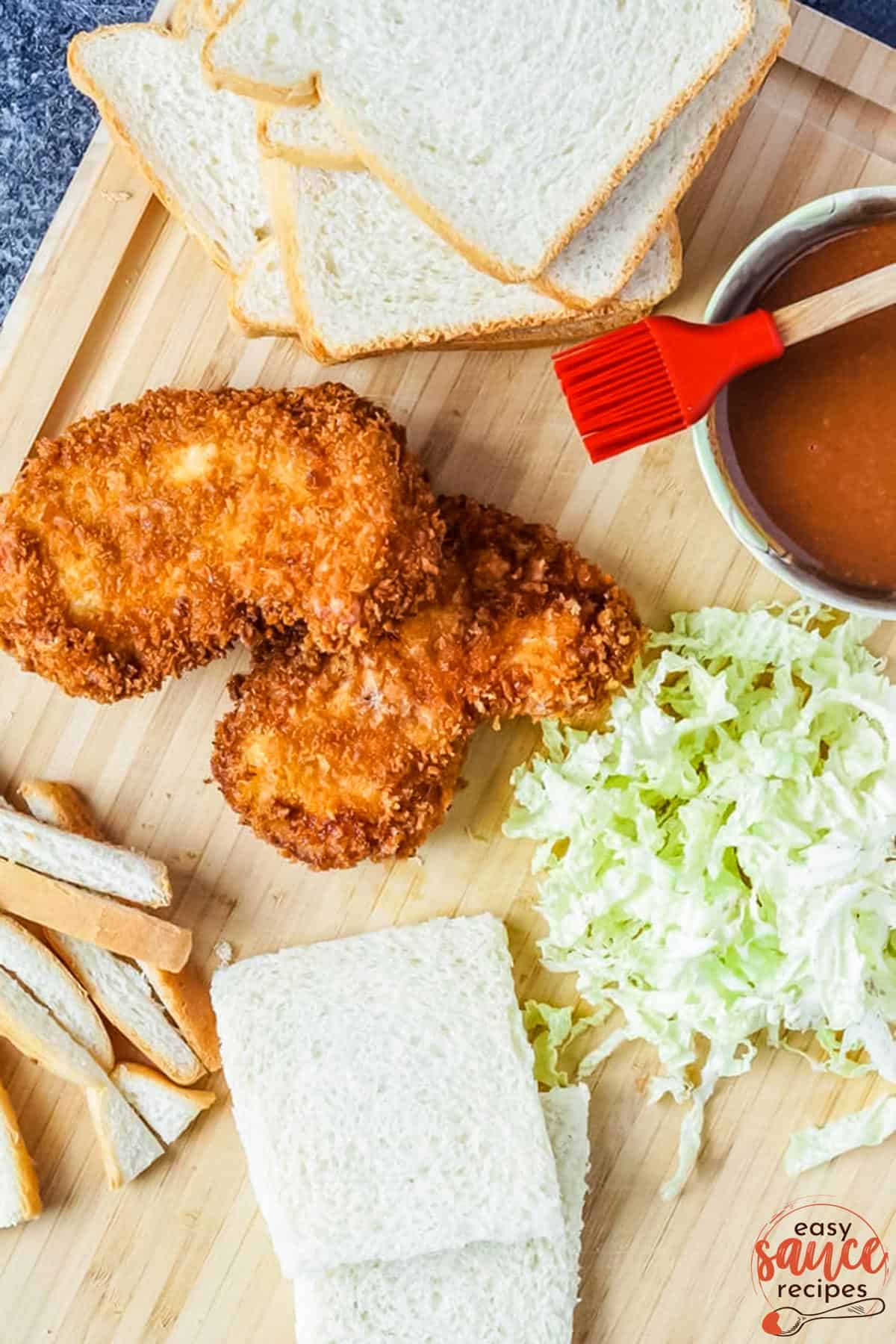 a cutting board with sliced white bread, pork cutlets and shredded cabbage next to a bowl of katsu sauce.