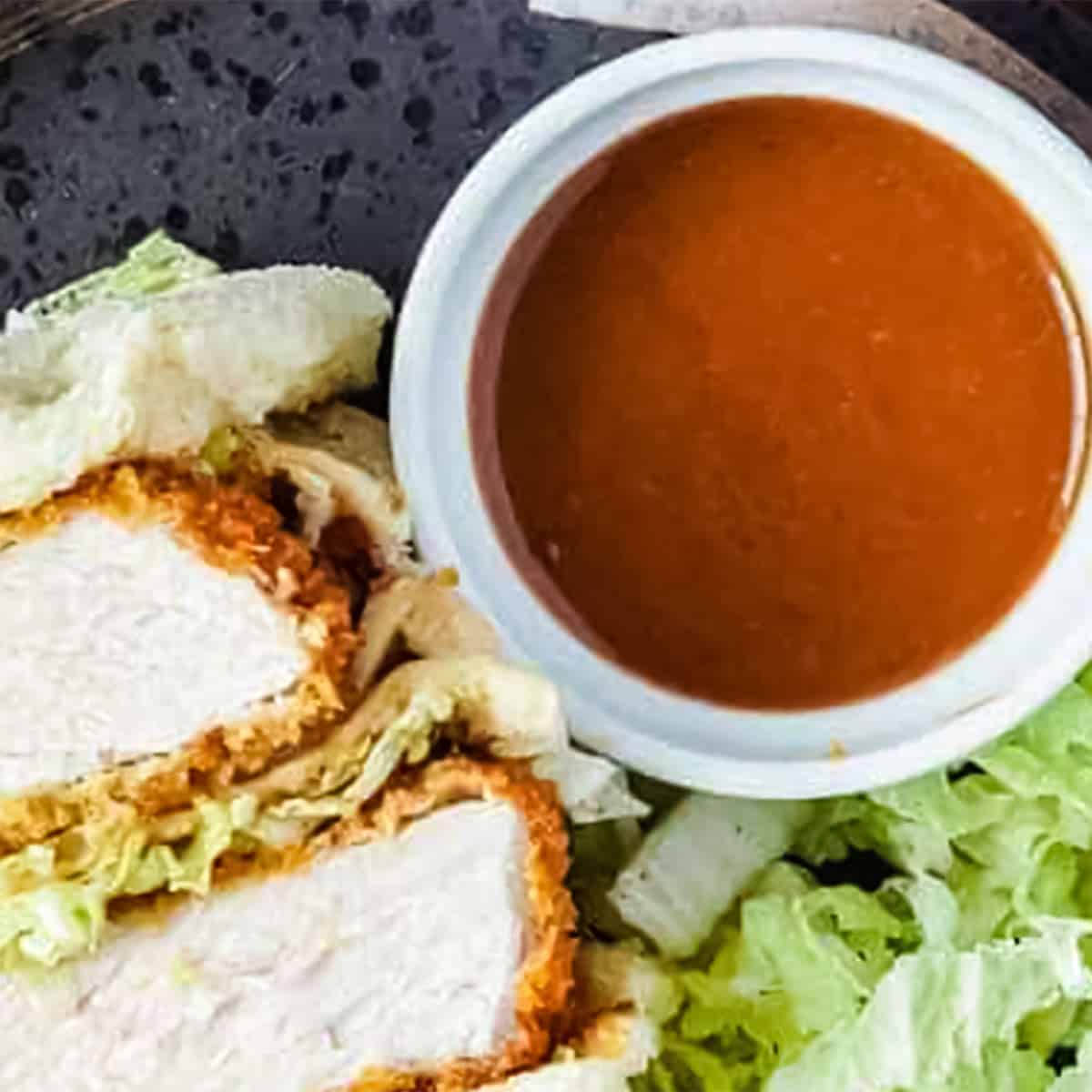 a bowl of katsu sauce, a pork cutlet sandwich and shredded cabbage on a dark plate.