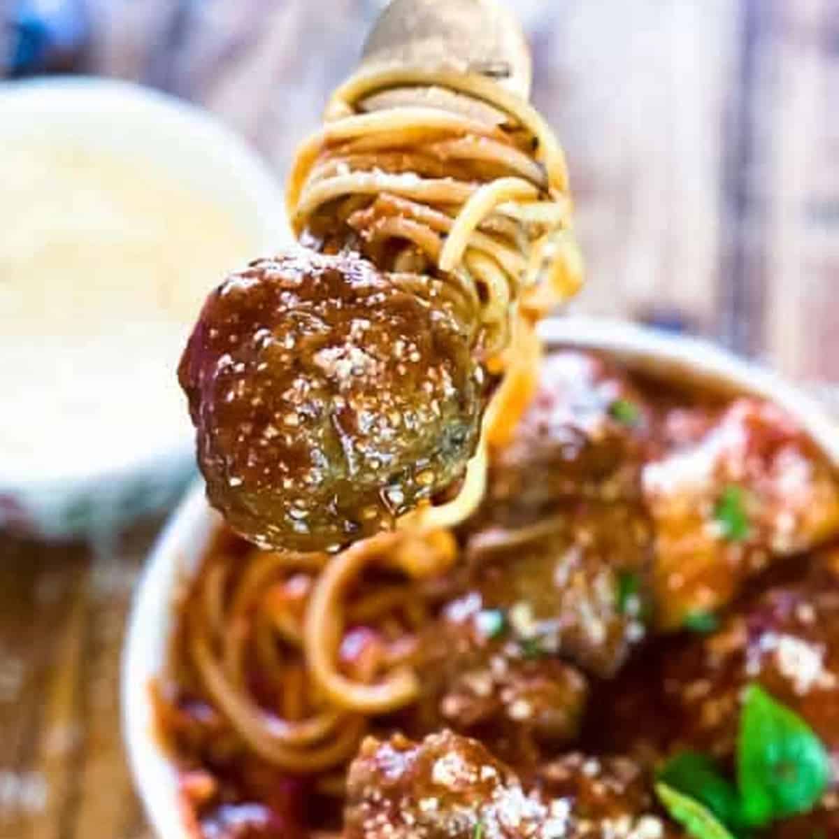 a meatball with spaghetti and sauce being lifted on a fork over a bowl of meatballs