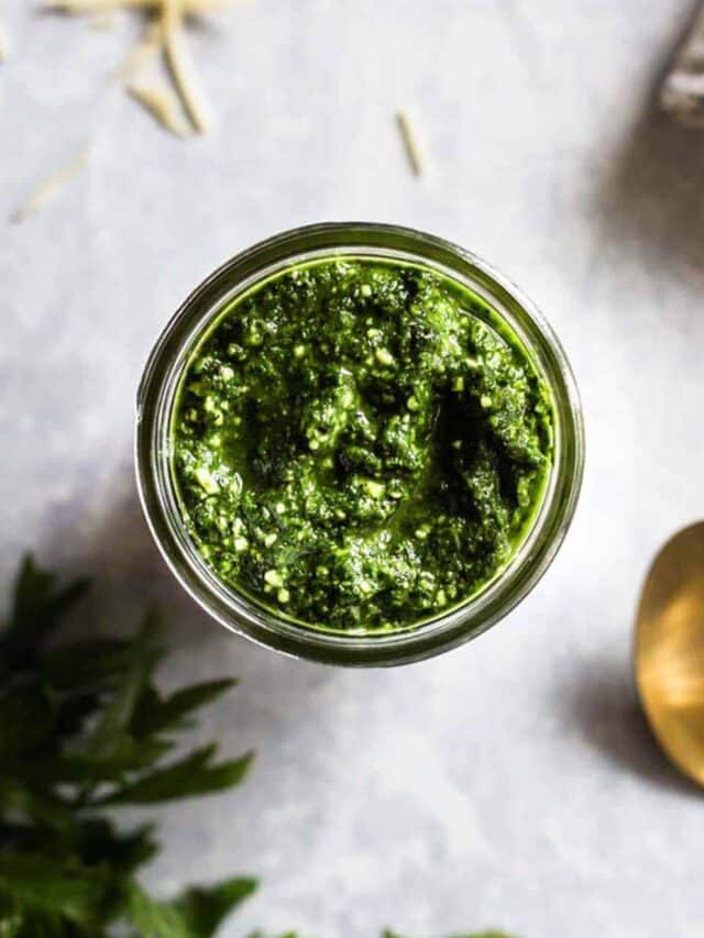 parsley pesto in a glass jar next to a spoon, parsley, and parmesan