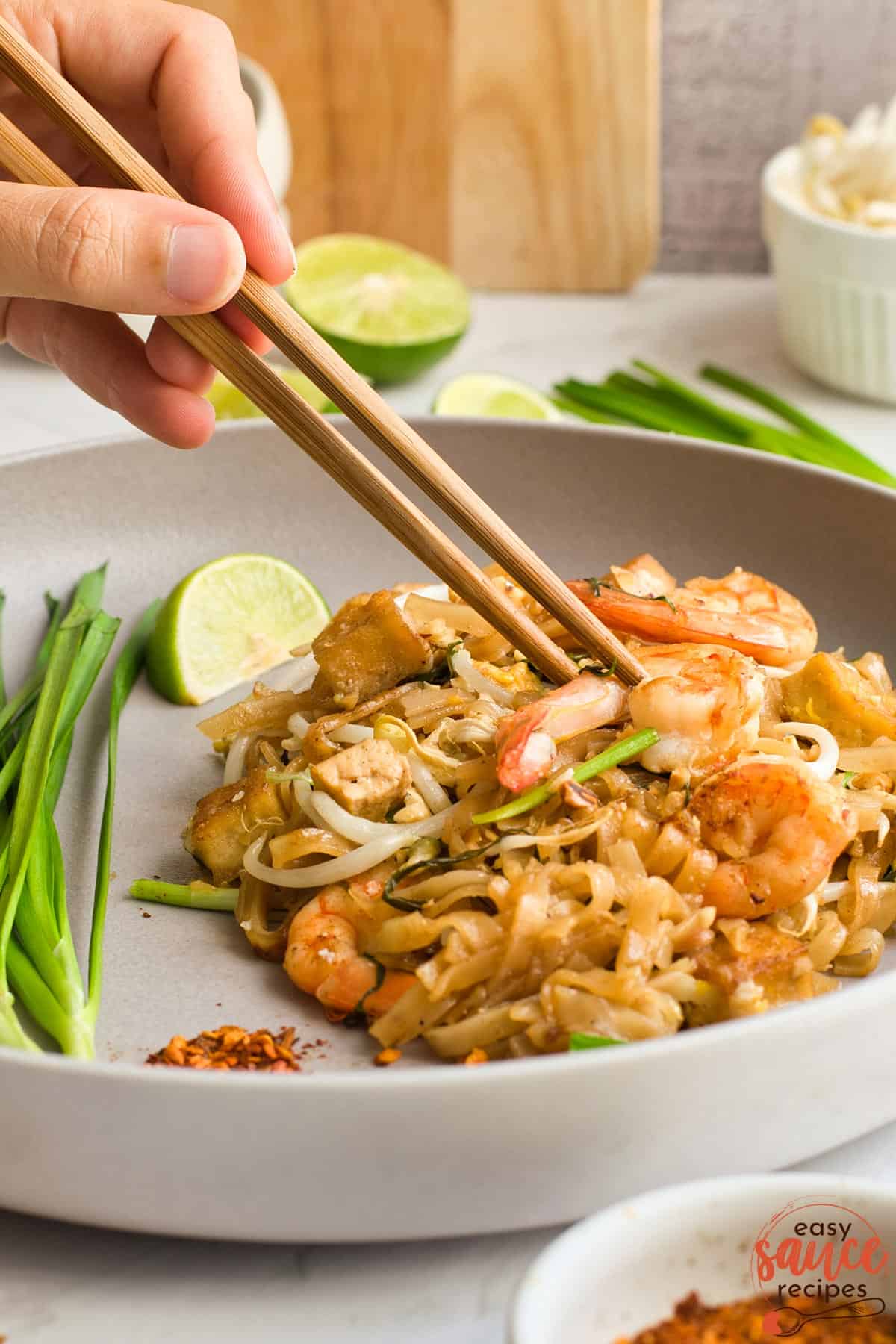 chopsticks taking a shrimp off of a dish of pad thai with added lime, chili, and green onions