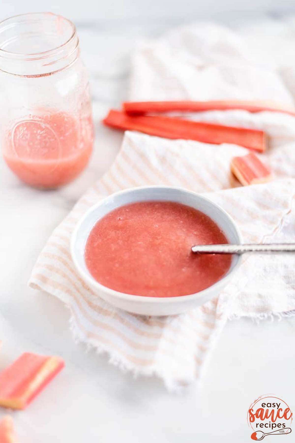 rhubarb sauce in a white bowl with a spoon next to a jar of sauce and fresh rhubarb