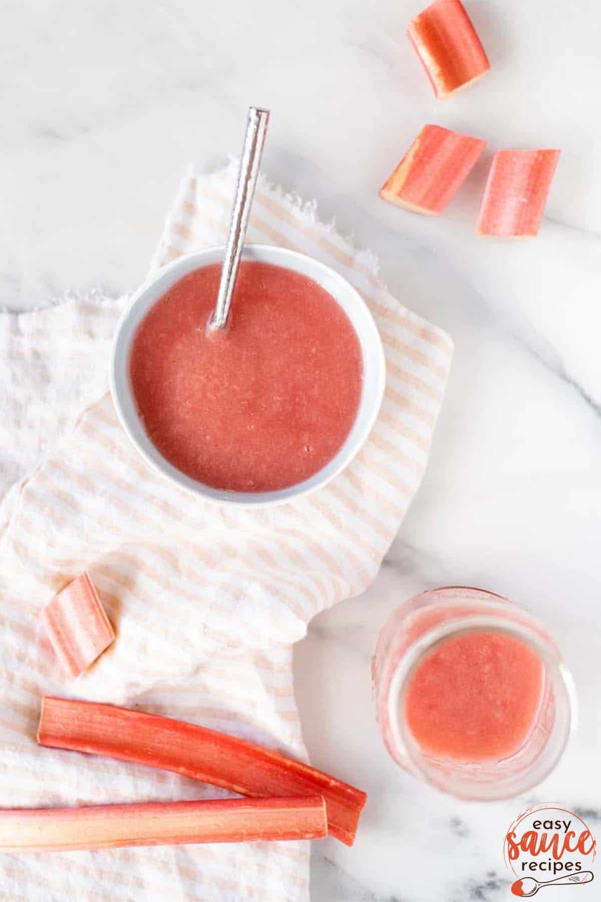 a bowl of rhubarb sauce with a spoon next to a jar of sauce and cut rhubarb